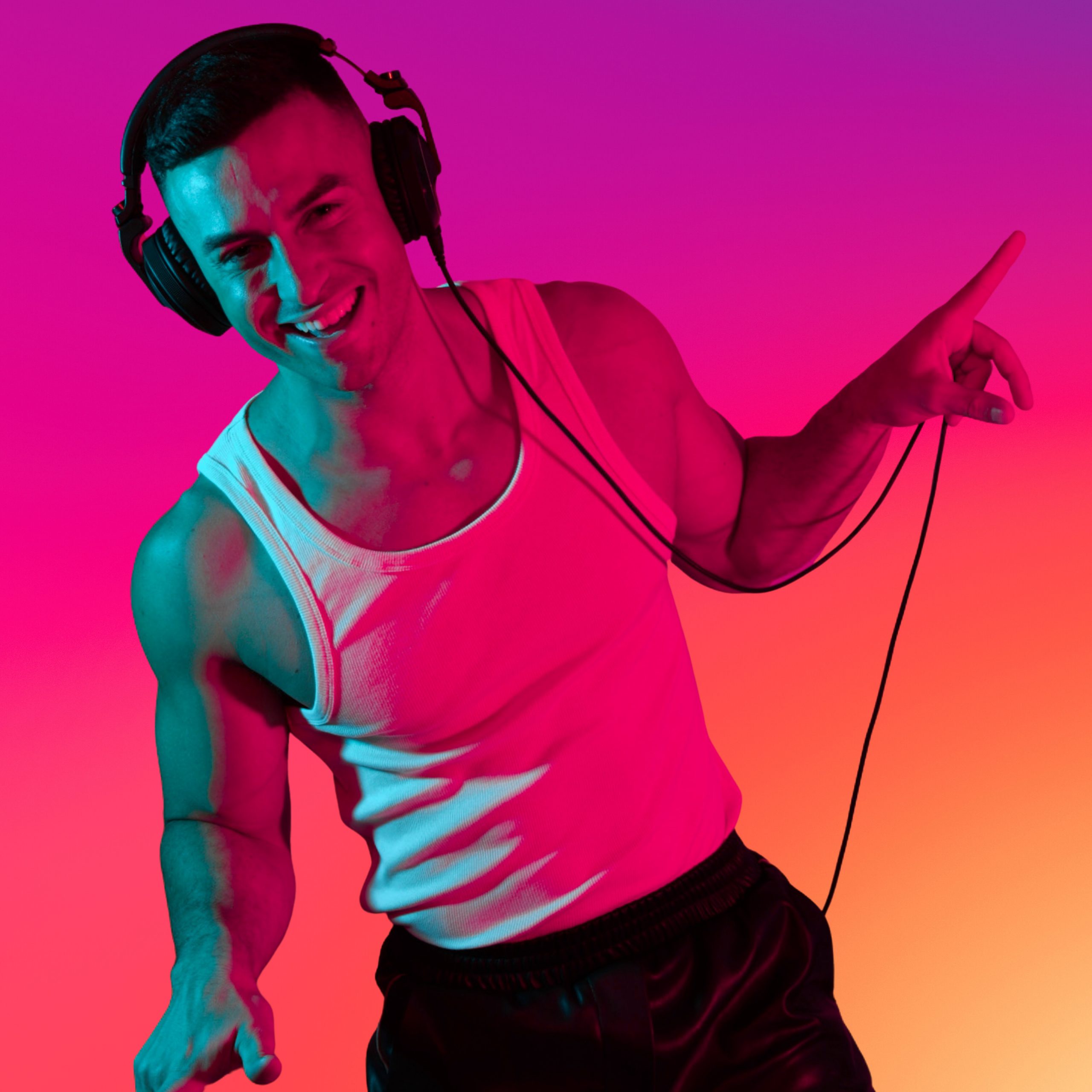 Maykel Moreira in headphones and a white tank top, smiling and pointing, lit against a pink and orange background